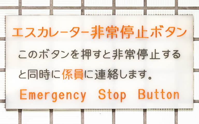 「エスカレーター非常停止ボタン このボタンを押すと非常停止すると同時に係員に連絡します Emergency Stop Button」と書かれたアクリル板の写真。文字は機械彫刻用標準書体。