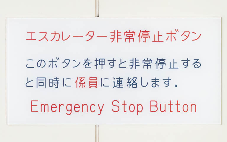 「エスカレーター非常停止ボタン このボタンを押すと非常停止すると同時に係員に連絡します Emergency Stop Button」と書かれたアクリル板の写真。文字は機械彫刻用標準書体。