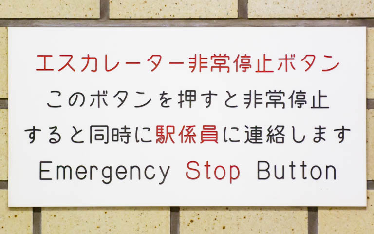 「エスカレーター非常停止ボタン このボタンを押すと非常停止すると同時に係員に連絡します Emergency Stop Button」と書かれたアクリル板の写真。文字は機械彫刻用標準書体ではない。