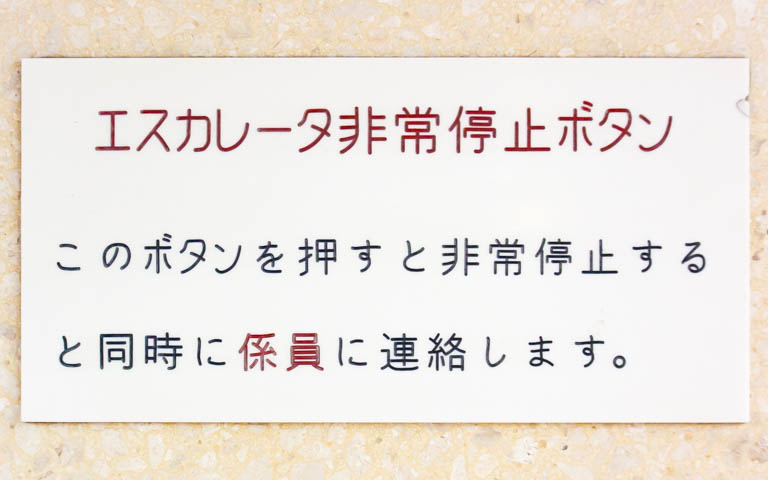 「エスカレーター非常停止ボタン このボタンを押すと非常停止すると同時に係員に連絡します。」と書かれたアクリル板の写真。文字は機械彫刻用標準書体ではない。