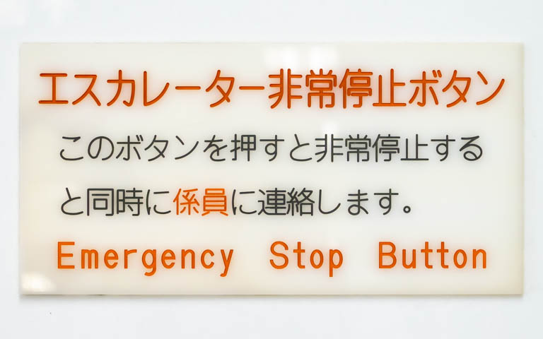 「エスカレーター非常停止ボタン このボタンを押すと非常停止すると同時に係員に連絡します。 Emergency Stop Button」と書かれたアクリル板の写真。文字は機械彫刻用標準書体ではない。