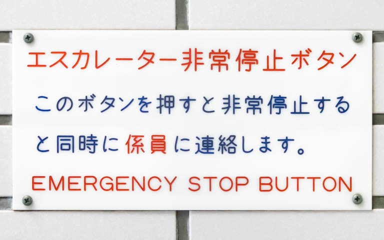 「エスカレーター非常停止ボタン このボタンを押すと非常停止すると同時に係員に連絡します。EMERGENCY STOP BUTTON」と書かれたアクリル板の写真。文字は漢字のみ機械彫刻用標準書体であり、仮名と英字は機械彫刻用標準書体ではない。