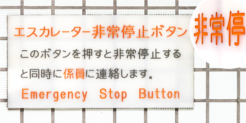「エスカレーター非常停止ボタン このボタンを押すと非常停止すると同時に係員に連絡します EMERGENCY STOP BUTTON」と書かれた乳白色アクリル板の写真。